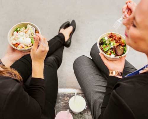 Two women seated outdoors, eating fresh salad bowls with mixed greens and toppings, accompanied by drinks.