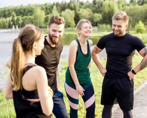 A group of four friends in activewear smiling and chatting outdoors near a lake, with a forested area in the background.