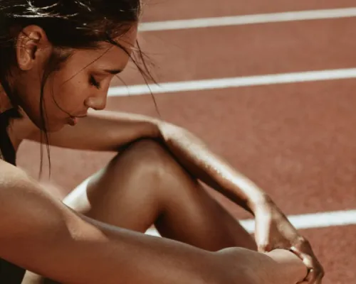 A runner in a black sports top and shorts crouches on a track, fastening her shoelace while leaning forward, with white lane lines in the background.