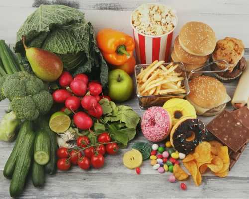 A variety of healthy vegetables and fruits next to fast food items like burgers, fries, donuts, and popcorn.