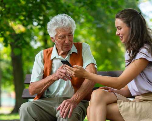 A young woman showing an elderly man how to use a smartphone while they sit on a park bench. The man has a continuous glucose monitor on his arm.