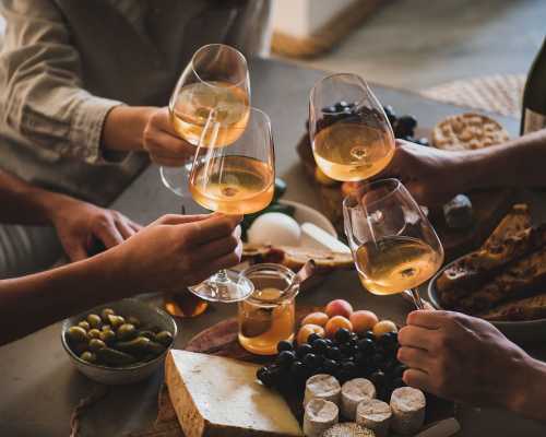 Friends toasting with white wine over a table of cheese, bread, grapes, and olives.