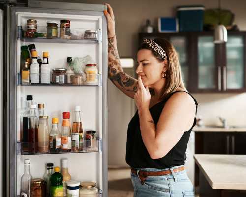 Femme debout devant un réfrigérateur ouvert, réfléchissant en regardant différents aliments et boissons à l'intérieur.  Woman standing in front of an open fridge, thoughtfully looking inside at various food items and drinks.