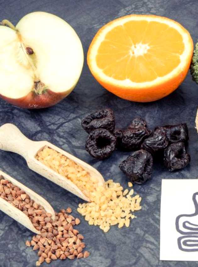 Fibre-rich foods including fruits like apples and oranges, vegetables like broccoli, whole grain bread, dried apricots, prunes, as well as cereals such as buckwheat and brown rice, placed next to an illustration of a stomach to represent potential discomforts related to the presence of FODMAPs.