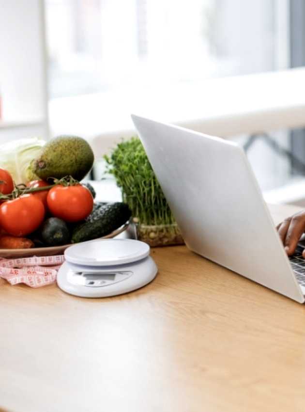 A nutritionist and Registered Dietitian in a white coat works on a laptop with fresh fruits, vegetables, and supplements on the table.