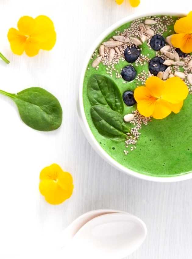 A vibrant green smoothie bowl topped with yellow edible flowers, fresh blueberries, chia seeds, sunflower seeds, and baby spinach leaves. The bowl is arranged on a light background with additional spinach leaves and yellow flowers scattered around for decoration.