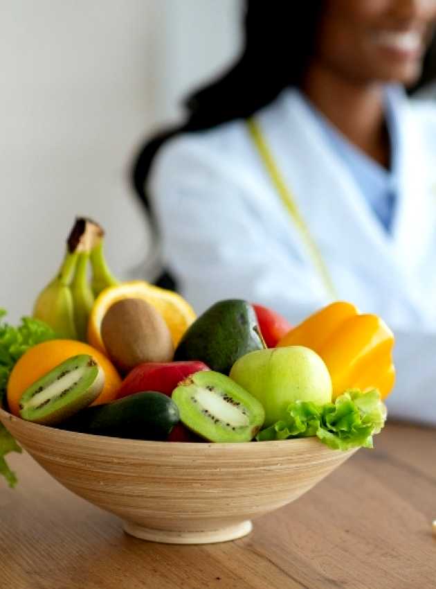 A bowl of fresh fruits, including kiwi, apple, and banana, with a glass of juice on a table, while a nutritionist works on a laptop in the background.