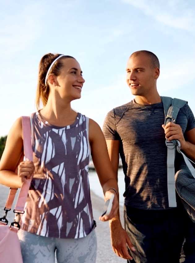 A smiling couple in activewear walks on a beach, each carrying a gym bag, with the sea and trees in the background.