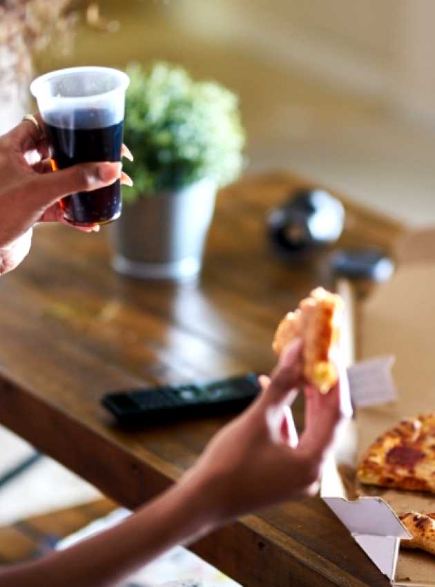 Person holding a slice of pepperoni pizza and a cup of soda, with a pizza box on a wooden table.