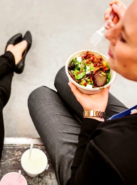 Two women seated outdoors, eating fresh salad bowls with mixed greens and toppings, accompanied by drinks.