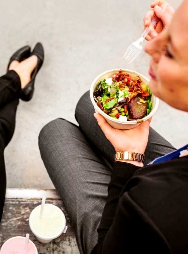 Two women seated outdoors, eating fresh salad bowls with mixed greens and toppings, accompanied by drinks.