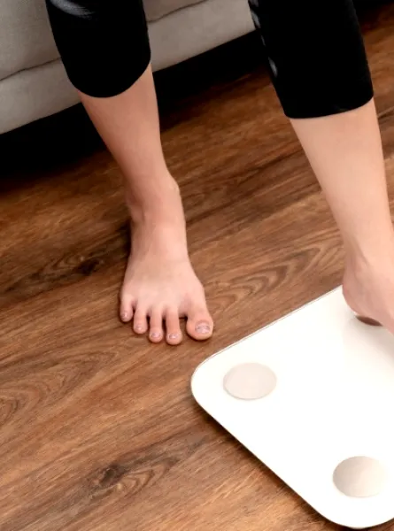 Person stepping on a white scale in a living room with pink dumbbells on the wooden floor nearby.