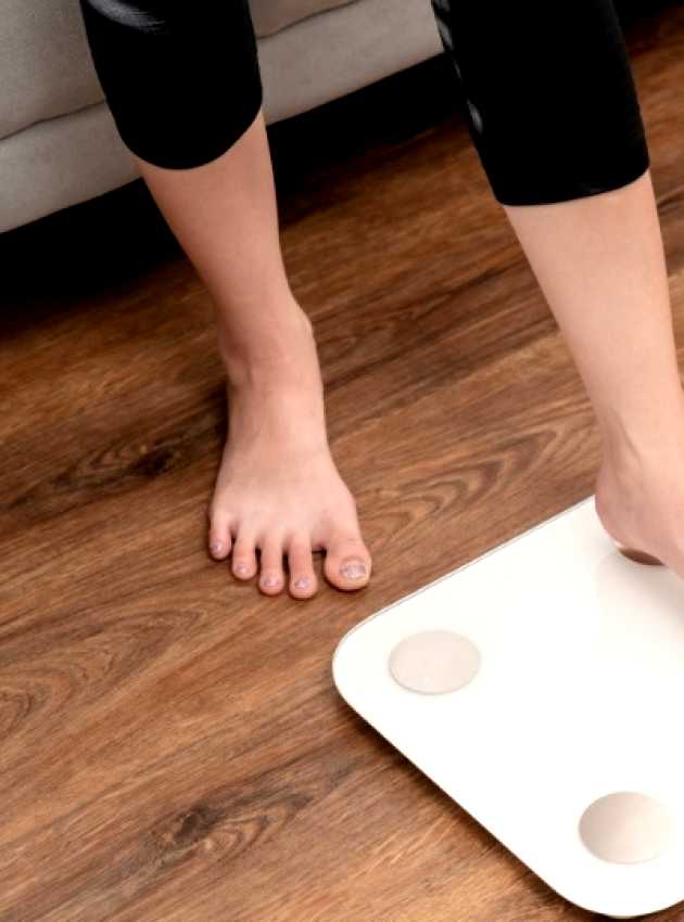 Person stepping on a white scale in a living room with pink dumbbells on the wooden floor nearby.