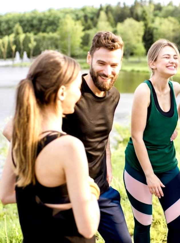 A group of four friends in activewear smiling and chatting outdoors near a lake, with a forested area in the background.