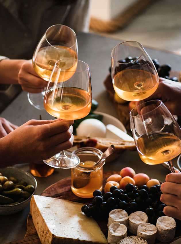 Friends toasting with white wine over a table of cheese, bread, grapes, and olives.