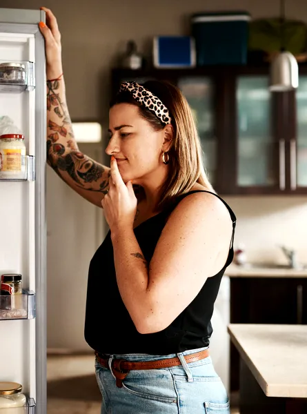 Femme debout devant un réfrigérateur ouvert, réfléchissant en regardant différents aliments et boissons à l'intérieur.  Woman standing in front of an open fridge, thoughtfully looking inside at various food items and drinks.