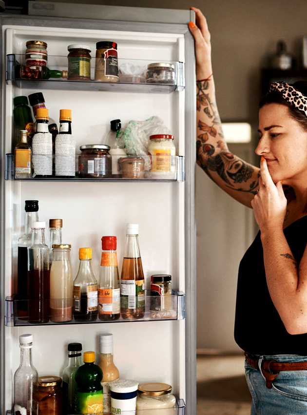 Femme debout devant un réfrigérateur ouvert, réfléchissant en regardant différents aliments et boissons à l'intérieur.  Woman standing in front of an open fridge, thoughtfully looking inside at various food items and drinks.