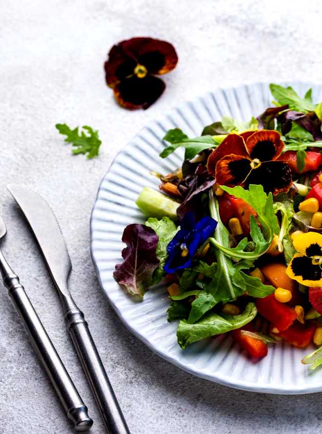 Une salade colorée garnie de fleurs comestibles, avec des légumes variés et des pensées violettes, jaunes et blanches - A colourful salad garnished with edible flowers, featuring various vegetables and purple, yellow, and white pansies